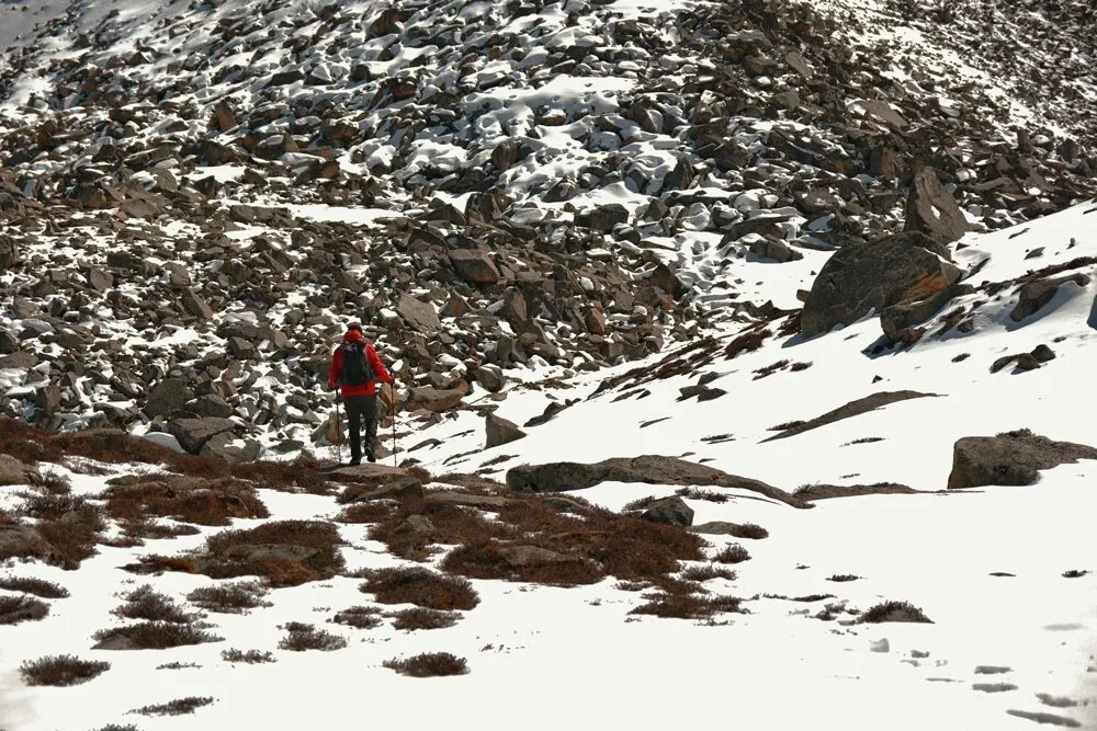A hiker in a red jacket trekking across a vast, snow-covered and rocky mountain landscape in Western Sichuan.