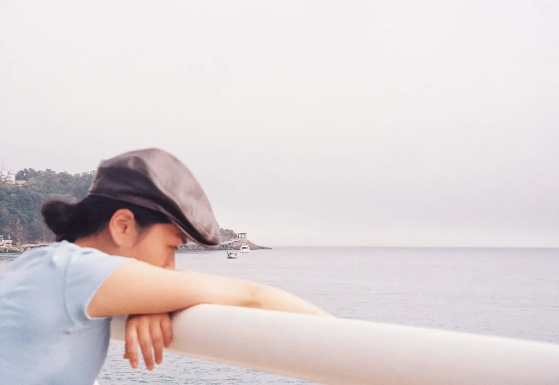 A person in a cap leans on a railing, looking out at the calm sea under an overcast sky.