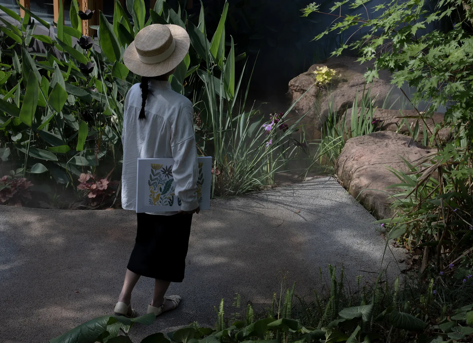 Embroidery artist wearing a straw hat and holding her artwork while walking on a path in a lush garden.