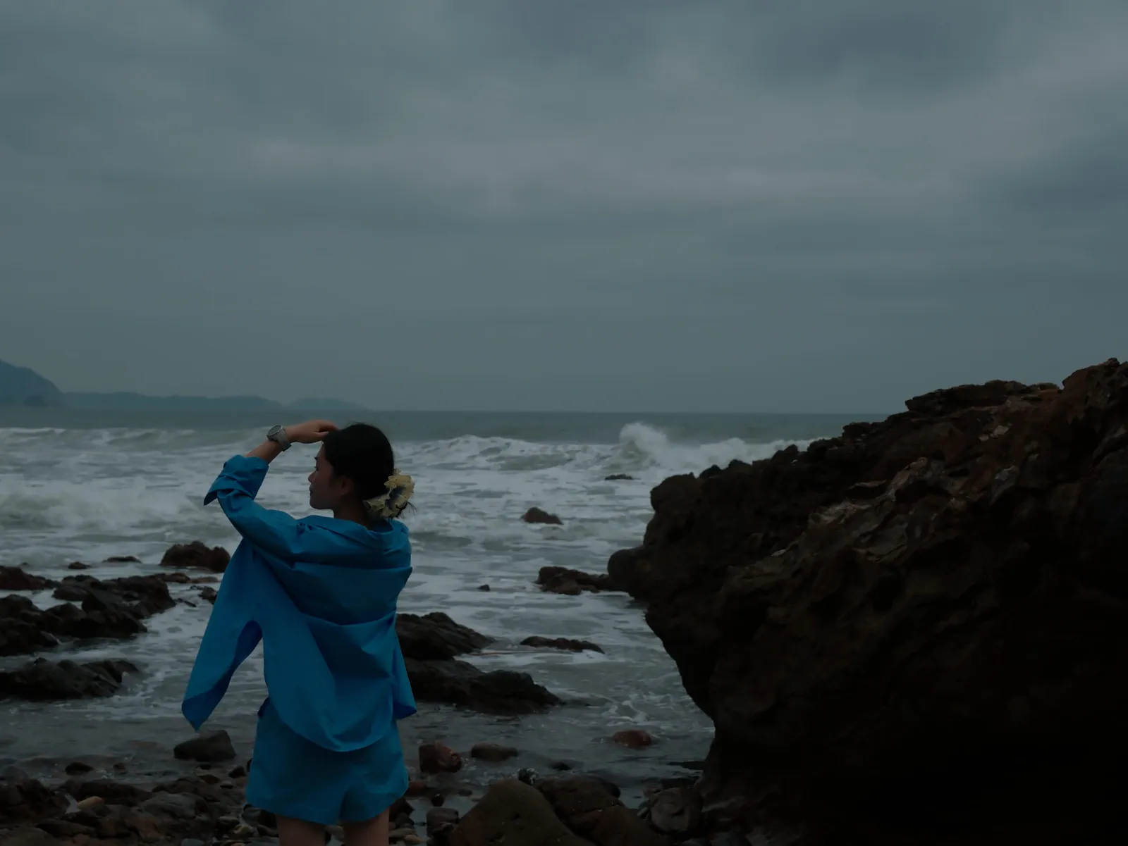 Lissyluo in a bright blue outfit on a rocky shore, looking out at the ocean under a moody, overcast sky.