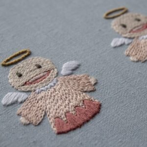 Close-up of a smiling embroidered angel, showing the texture of the satin stitch and thread details on its face and body.