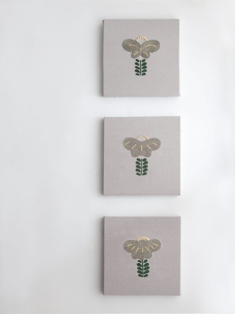 The hand-embroidered triptych "Botanical Observation No. 5" by lissyluo, displayed vertically on a white wall. Each of the three panels features an identical stylized grey and green floral motif on textured grey canvas.