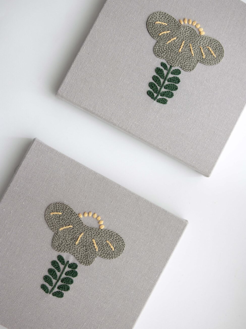 Overhead view of two panels from the "Botanical Observation No. 5" triptych, showing the repeated embroidered floral motif on grey canvas.