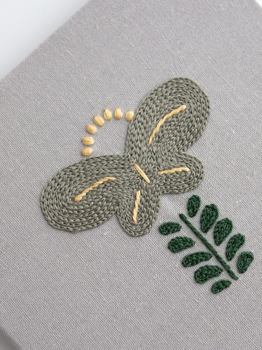 Close-up of a single panel from "Botanical Observation No. 5," showing the detailed texture of the grey chain stitch flower and green satin stitch leaves on grey canvas.