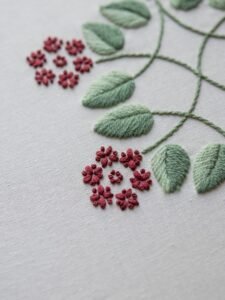 Detailed macro shot of a botanical hand embroidery, focusing on the texture of the red flower clusters and green satin-stitched leaves on linen.