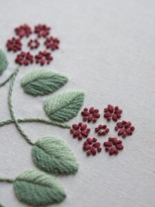 Extreme close-up of embroidery details, showing the texture of the sage green satin stitch leaves and the tiny red French knot flowers.