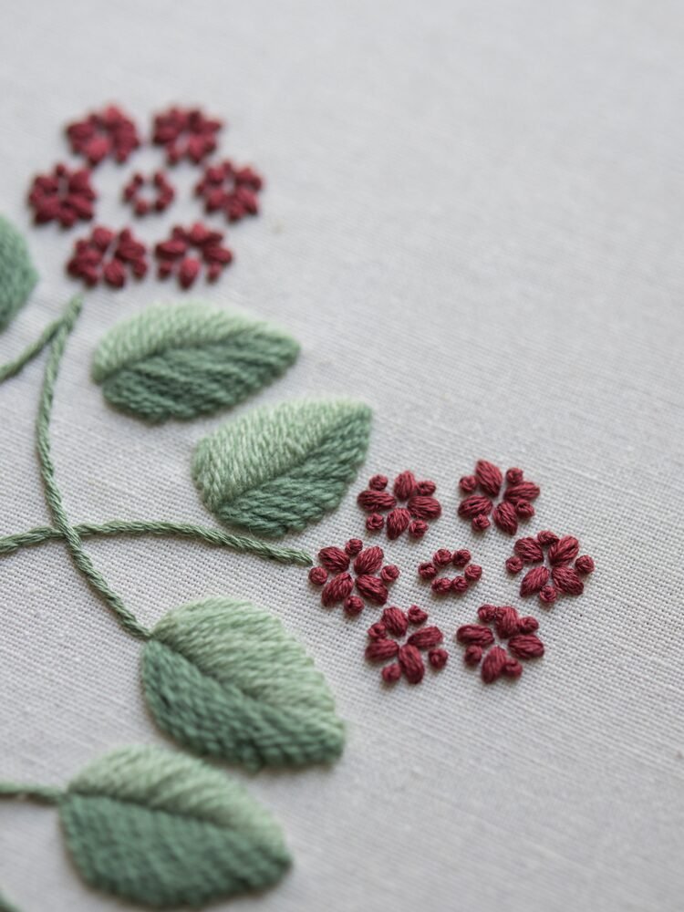 Extreme close-up of embroidery details, showing the texture of the sage green satin stitch leaves and the tiny red French knot flowers.