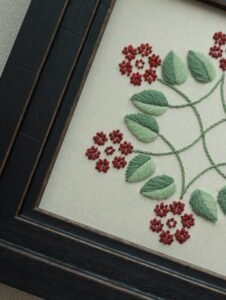 Angled detail shot of hand-stitched red flowers and green leaves, highlighting the intricate thread work within the black wooden frame.