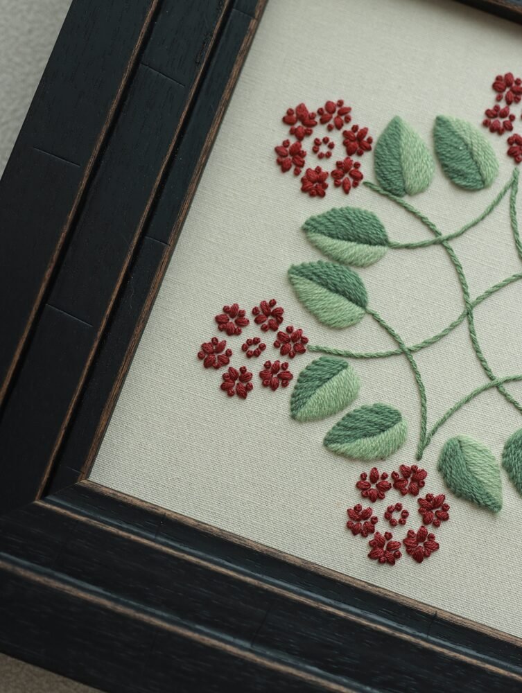 Angled detail shot of hand-stitched red flowers and green leaves, highlighting the intricate thread work within the black wooden frame.