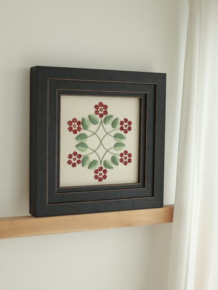 Framed botanical hand embroidery by lissyluo on a wooden shelf. The piece features a symmetrical pattern of red flowers and sage green leaves in a black wood frame.