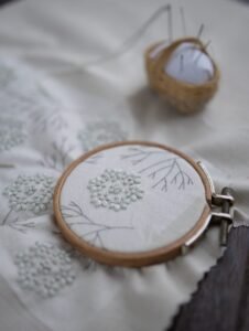 A work-in-progress shot of a botanical hand embroidery in a wooden hoop, showing delicate white flower clusters on natural linen. A small pincushion is blurred in the background.