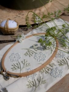 A still life composition featuring the "Botanical Observation No. 7" embroidery in a hoop, styled with fresh wildflower cuttings and a woven pincushion.