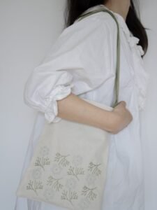 Lissyluo is wearing a white blouse holds a natural canvas tote bag with a sage green strap. The bag is hand-embroidered with a delicate pattern of white wildflower clusters and sage green stems.`