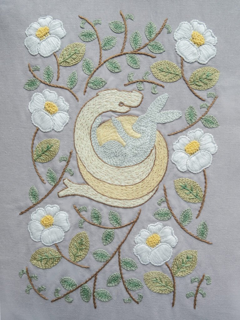 Completed hand embroidery of 'The Year of Snake,' featuring a central coiled snake and rabbit motif surrounded by a border of white flowers and green leaves on grey fabric.