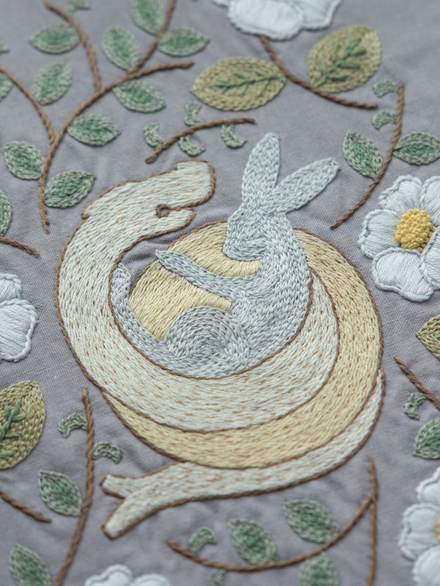 Close-up detail of 'The Year of Snake' hand embroidery, showing a cream-colored snake coiled around a light blue rabbit, surrounded by white flowers.
