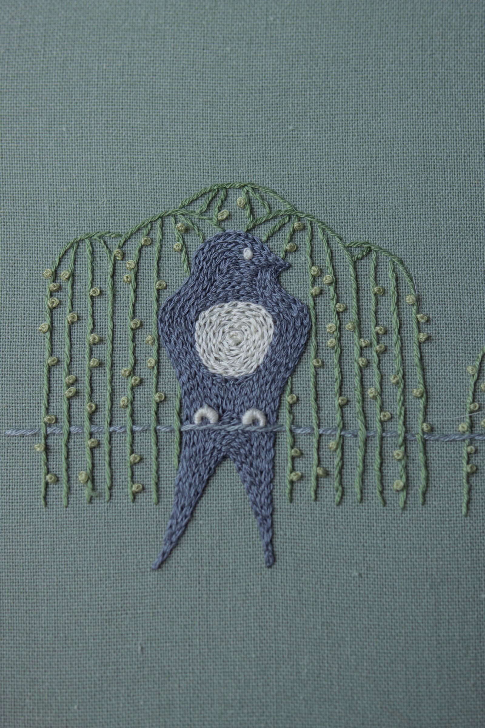 Close-up of a stylized bird hand-embroidered by lissyluo. The blue-gray bird, with a white spiral-stitched belly, sits under green weeping willow branches on sage green fabric.