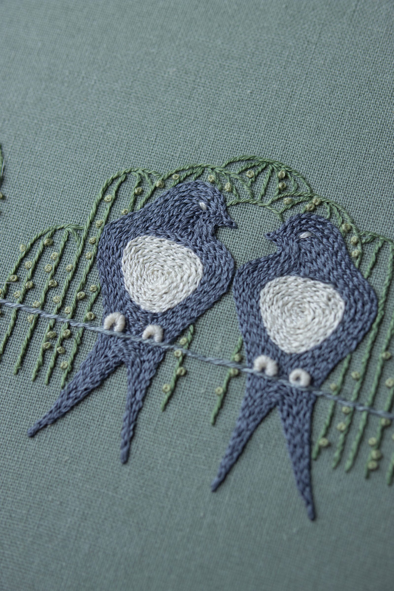 A detailed close-up of two embroidered birds facing each other by lissyluo. The pair of blue-gray birds sit on a stitched line, framed by delicate green willow branches.