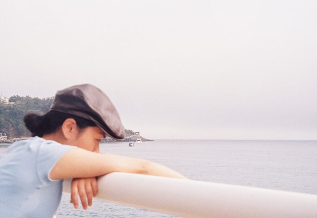 A Whisper Across Time: For the Art That Waits for Its Listener A person in a cap leans on a railing, looking out at the calm sea under an overcast sky.