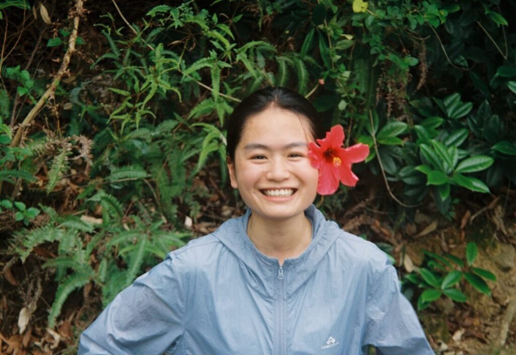 lissyluo is smiling with a red hibiscus flower tucked behind her ear, standing in front of green foliage.