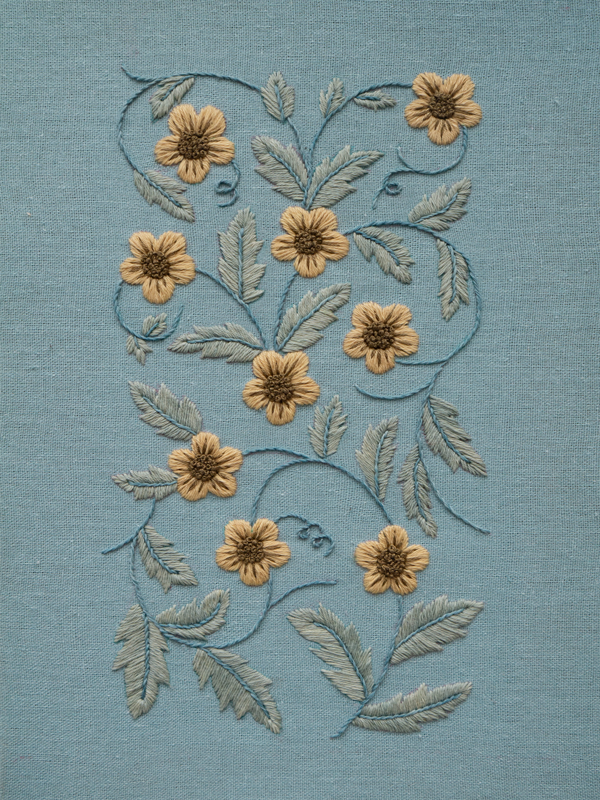Hand-embroidered floral vine with golden flowers and sage-colored leaves on a textured blue fabric, showing detailed satin and stem stitches.