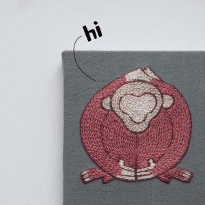 A detailed close-up of a single monkey motif from the 'Embracing Loneliness' hand embroidery. The pink and cream figure is stitched onto grey fabric that is neatly stretched and mounted for display.