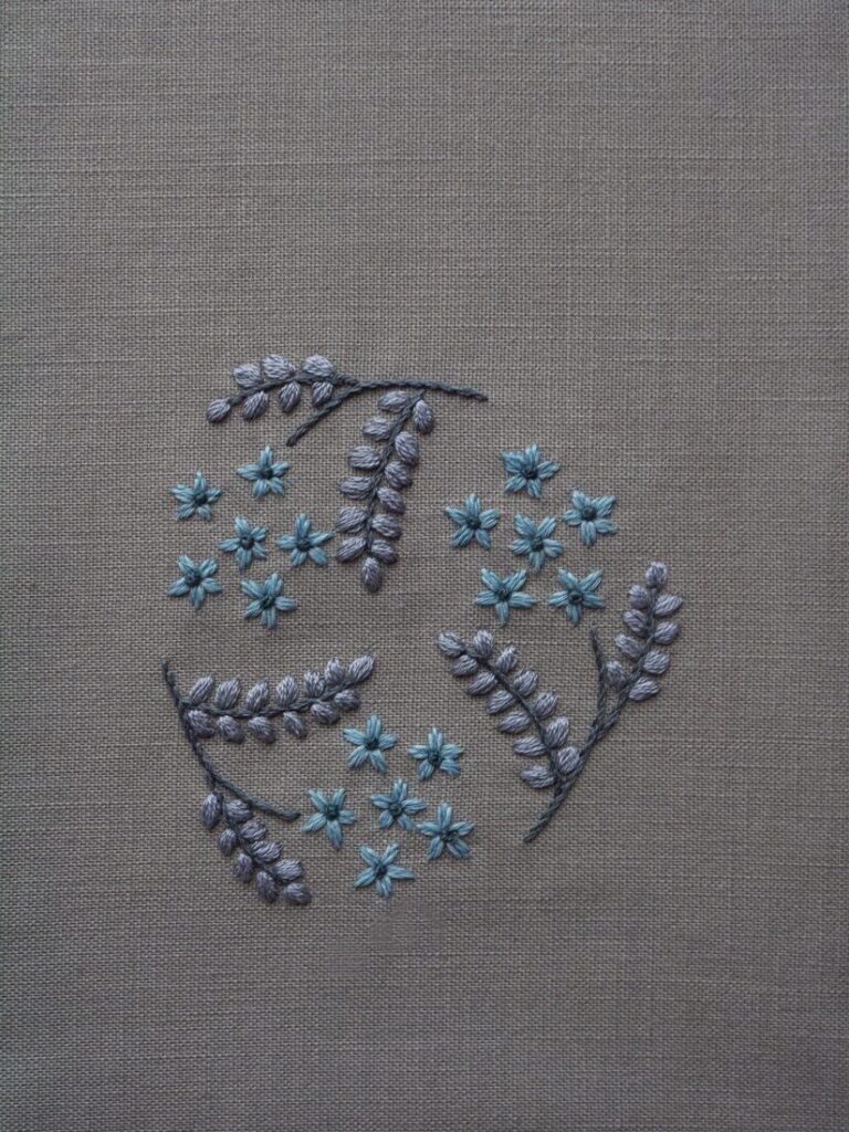 Botanical Observation No. 3 by lissyluo, a hand-embroidered artwork with a circular arrangement of grey leaves and dusty blue flowers on neutral linen fabric.