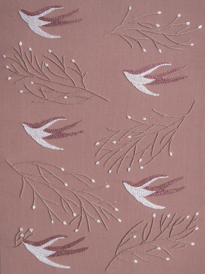 Hand-embroidered pattern of swallows in white and dusty rose thread, flying amongst budding branches on a mauve fabric.