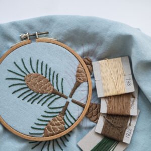 Work in progress shot of the "Pine" hand embroidery, showing the pinecone design in a wooden hoop next to bobbins of brown and green thread.