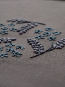 Angled view of Botanical Observation No. 3, a hand embroidery by lissyluo featuring dusty blue flowers and grey leaves on a taupe linen background.