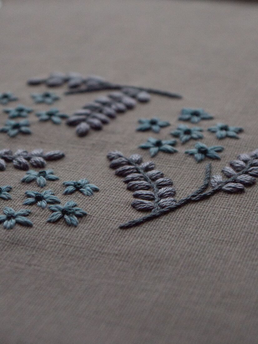 Angled view of Botanical Observation No. 3, a hand embroidery by lissyluo featuring dusty blue flowers and grey leaves on a taupe linen background.