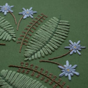 Detailed macro shot of botanical hand embroidery. Raised satin stitch fern leaves and French knot blue flowers create a 3D texture on the olive green canvas.