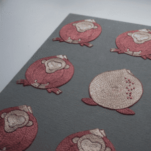 An angled perspective of the finished 'Embracing Loneliness' hand embroidery, emphasizing the raised texture and detail of the stitched figures on the grey fabric.