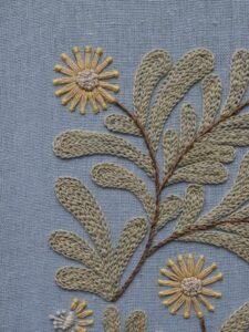 A top-down close-up of a hand-embroidered branch with stylized sage green leaves and a yellow flower on blue linen.