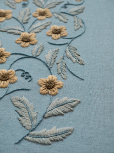 Angled close-up of a hand-embroidered floral vine with golden flowers and sage-colored leaves on a blue linen fabric.
