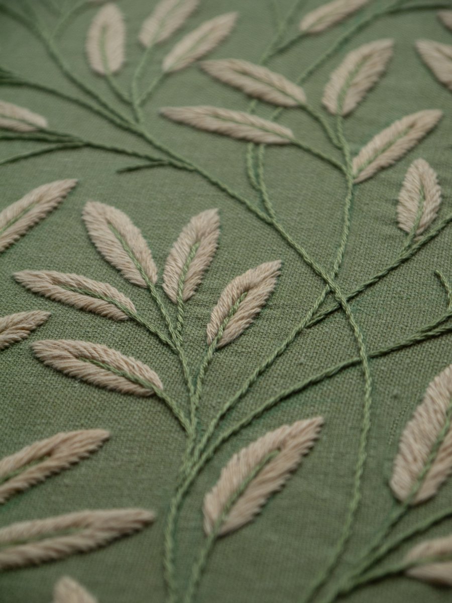 Detailed angled view of botanical hand embroidery, showing the texture of cream-colored leaf stitches on sage green fabric.