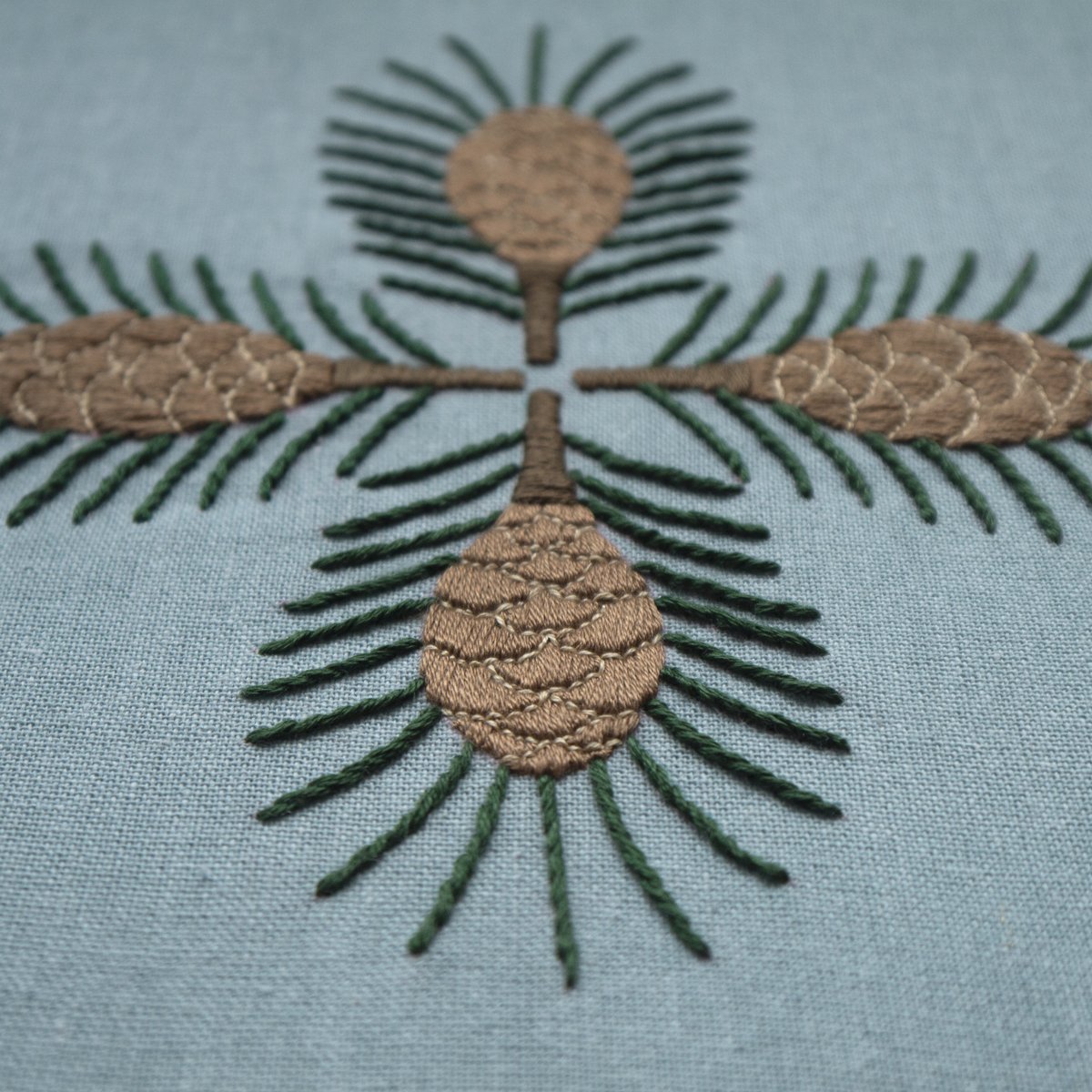 Detail shot of the "Pine" embroidery, focusing on the texture of the satin-stitched pinecones and straight-stitched needles on the blue fabric.