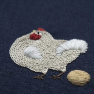 Close-up of a hand-embroidered chicken on navy blue fabric. The chicken's body is created with off-white chain stitch, its tail with fluffy white satin stitch, and it stands beside a golden-yellow embroidered egg.