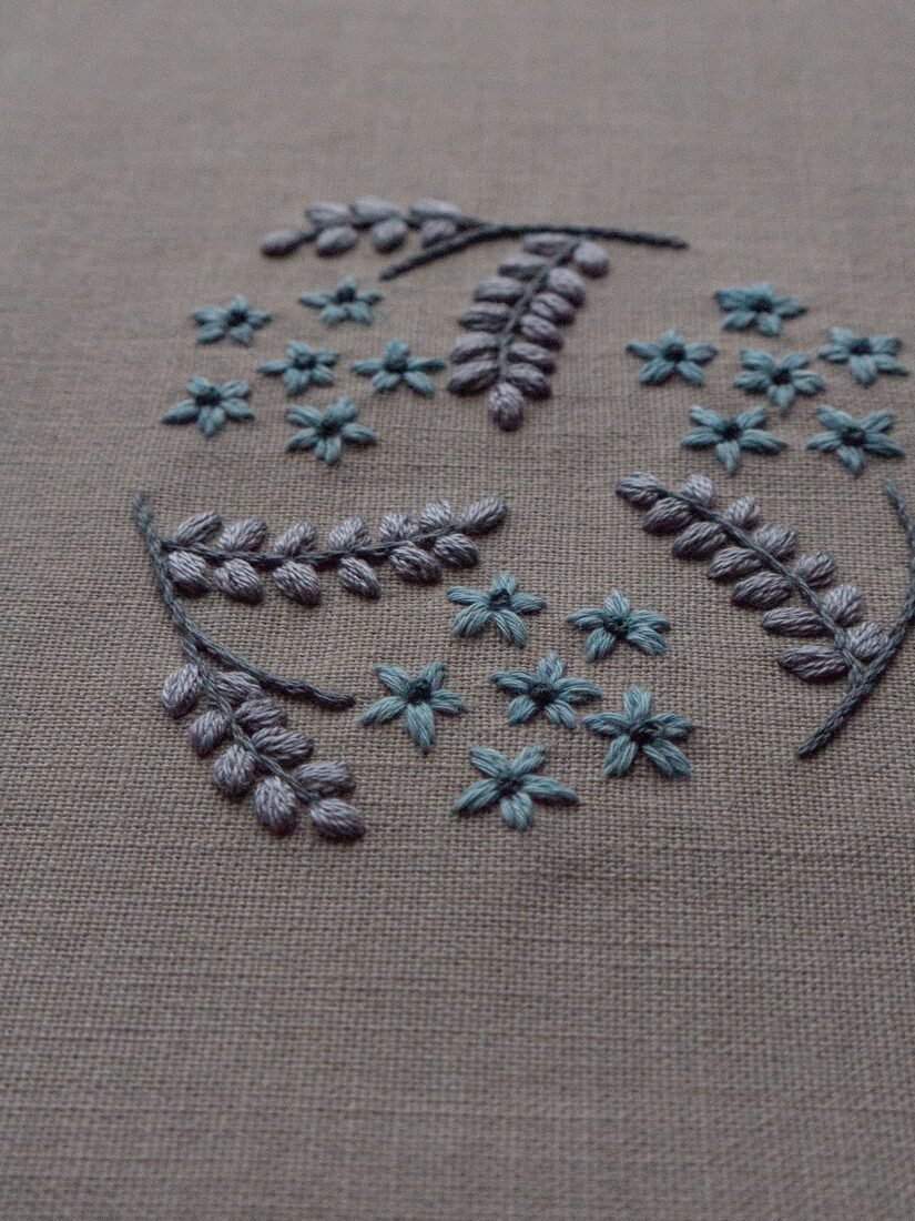 A detailed close-up shot of lissyluo's embroidery, highlighting the texture of the grey satin-stitched leaves and small blue flowers.
