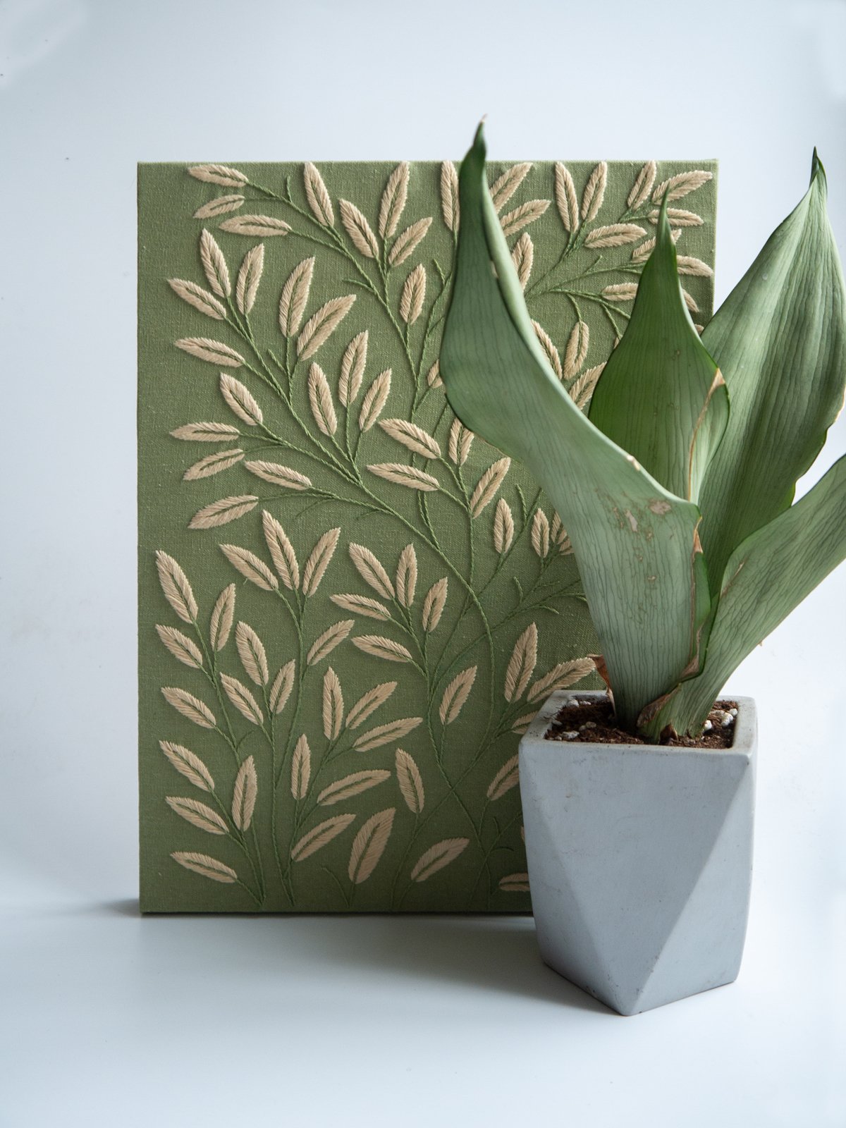 Finished botanical leaf embroidery on a sage green canvas, displayed next to a live snake plant in a geometric concrete pot.