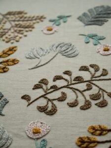 Angled detail shot of lissyluo's embroidery, highlighting the texture of a brown leafy branch and other botanical motifs on neutral fabric.