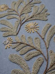 Detailed, angled close-up of hand embroidery showing the texture of sage green leaves and a yellow flower stitched on dusty blue fabric.