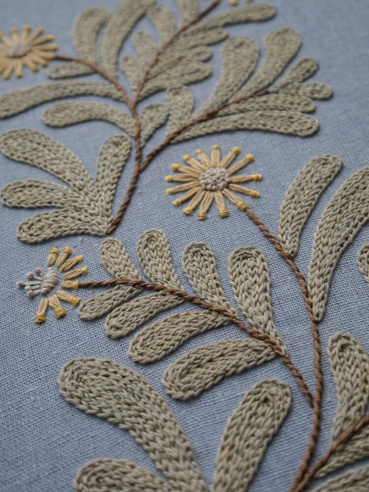 Detailed, angled close-up of hand embroidery showing the texture of sage green leaves and a yellow flower stitched on dusty blue fabric.