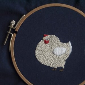 `A hand-embroidered plump chicken with an off-white body and red comb, stitched onto navy blue fabric and secured in a wooden embroidery hoop.`