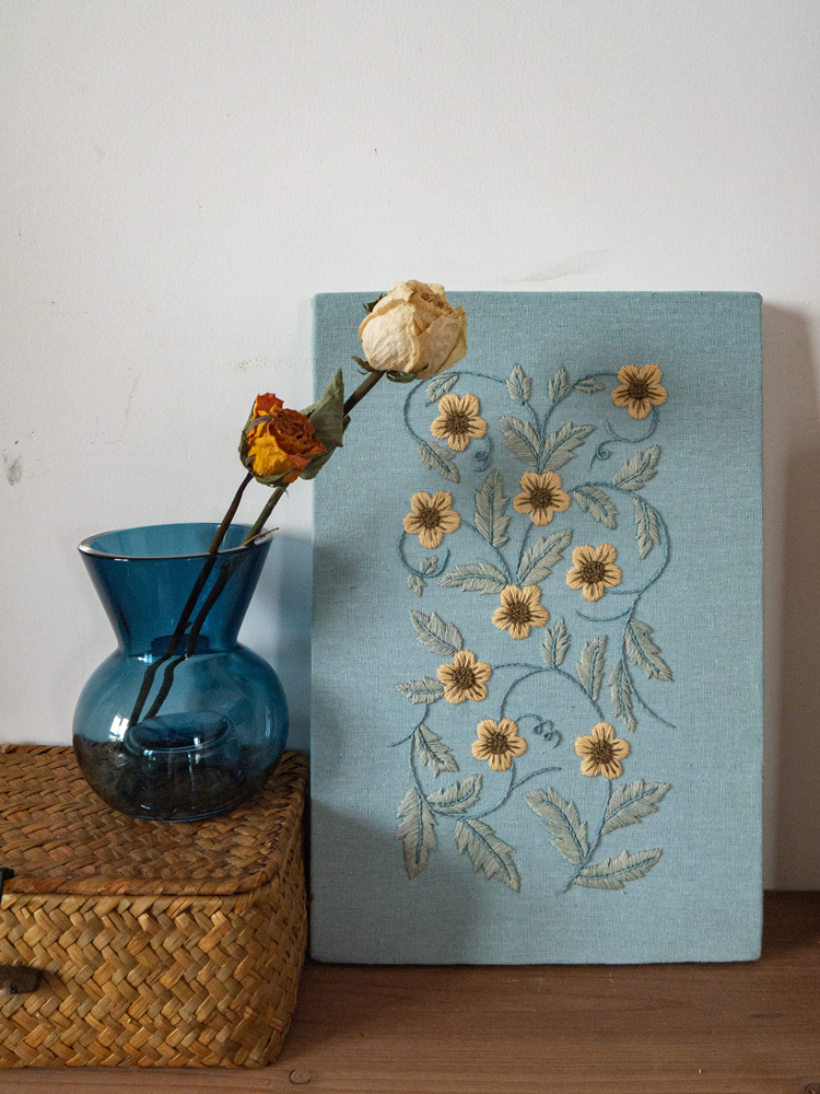 The mounted hand-embroidered floral art displayed leaning against a wall, styled with a blue glass vase of dried roses on a woven basket.