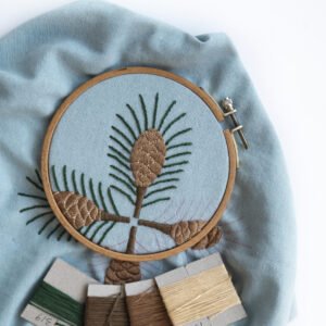 A flat lay of the "Pine" embroidery project, with the blue fabric in a hoop and the corresponding brown, green, and tan wool threads arranged nearby.