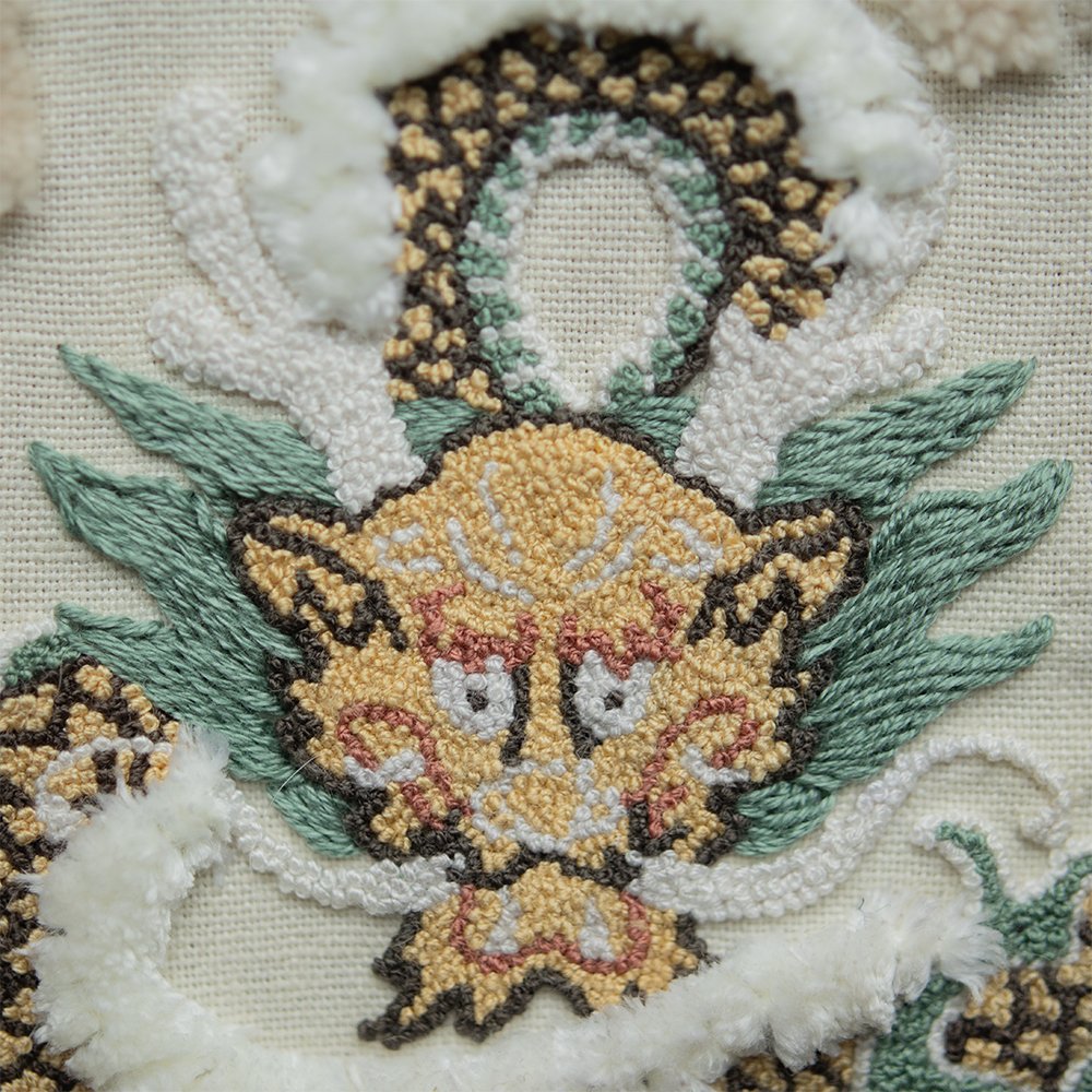 A macro shot of a hand-embroidered Chinese dragon's face, showing the textured punch needle stitches of its yellow face and white horns, contrasted with green satin stitch whiskers.