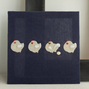 `Finished embroidery artwork of four chickens on a navy blue stretched canvas, displayed leaning against a black picture frame.