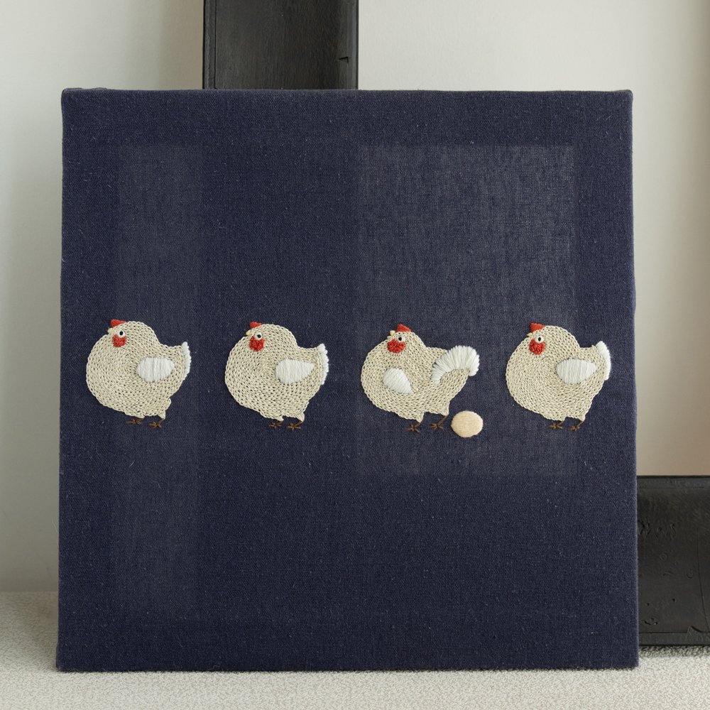 `Finished embroidery artwork of four chickens on a navy blue stretched canvas, displayed leaning against a black picture frame.