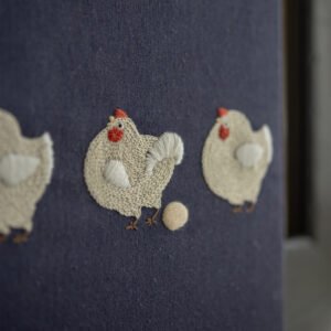 `Angled close-up of hand-embroidered chickens on navy blue canvas, focusing on the chicken that has just laid an egg.`