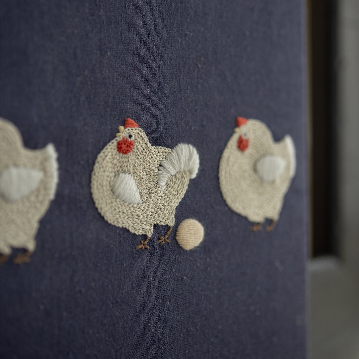 `Angled close-up of hand-embroidered chickens on navy blue canvas, focusing on the chicken that has just laid an egg.`